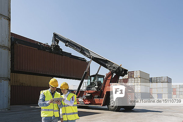 Arbeiter mit Tablette vor einem Kran mit Frachtcontainer auf einem Industriegelände