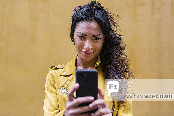 Porträt einer jungen Frau in gelber Lederjacke  die einen Selfie trägt  gelbe Wand im Hintergrund