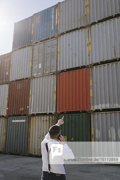 Rückansicht eines Managers  der vor Frachtcontainern auf einem Industriegelände per Handy telefoniert
