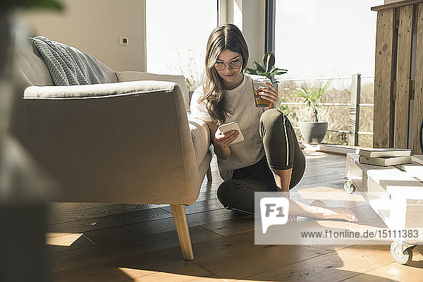 Junge Frau sitzt zu Hause mit Getränk und Handy auf dem Boden