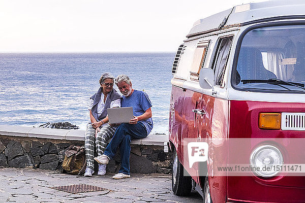 Älteres Ehepaar reist in einem Oldtimer-Lieferwagen  benutzt einen Laptop und sitzt auf einer Mauer am Meer