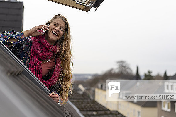 Glückliche junge Frau am Handy  die sich aus dem Dachfenster lehnt