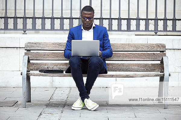 Young businessman wearing blue suit jacket sitting on bench and using laptop