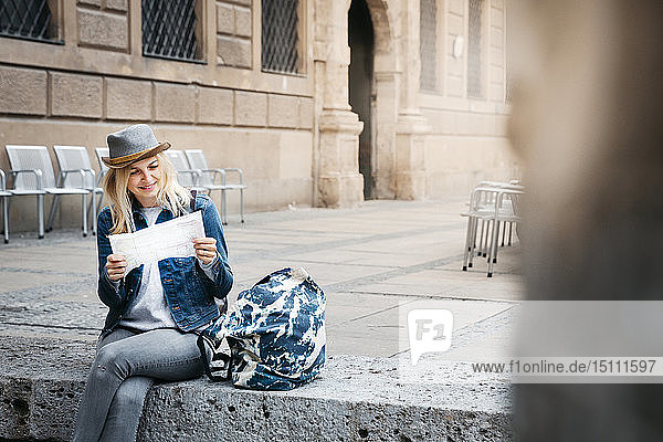 Smiling blond woman with baggage sitting on curb looking at map