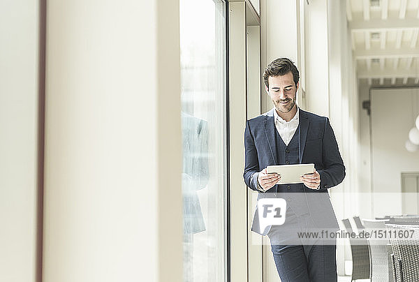 Junger Geschäftsmann steht im Bürogebäude  benutzt digitales Tablett  lehnt am Fenster