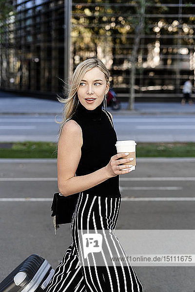 Porträt einer lächelnden blonden Frau mit Kaffee zum Mitnehmen und Rollgepäck