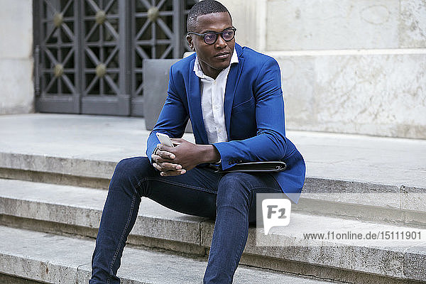 Junger Geschäftsmann in blauer Anzugjacke  sitzt auf einer Stufe und hält ein Smartphone