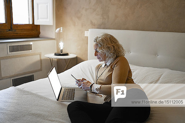 Reife Geschäftsfrau  die mit einem Laptop auf dem Bett liegt und ein Mobiltelefon benutzt