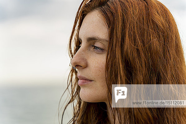 Porträt einer nachdenklichen rothaarigen jungen Frau mit Nasenpiercing vor dem Meer bei Sonnenuntergang