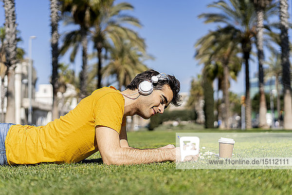 Spain  Barcelona  man lying on lawn in the city with headphones and notebook