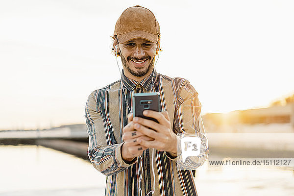 Portrait of smiling man wearing baseball cap listening music with earphones and smartphone at sunset