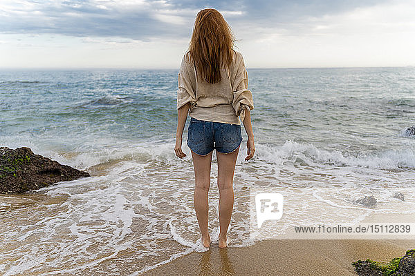 Back view of redheaded young woman standing on the beach looking to the sea