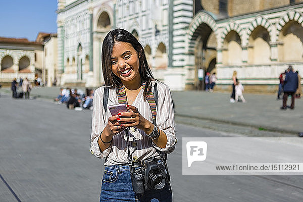 Italien  Florenz  Porträt eines glücklichen jungen Touristen  der mit Kamera und Rucksack auf sein Handy schaut