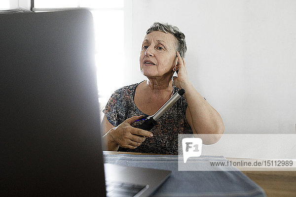 Ältere Frau sitzt zu Hause am Tisch mit Laptop und macht sich die Haare