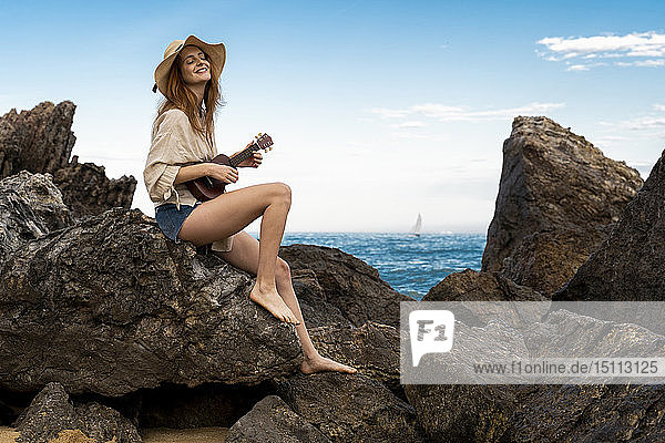 Glückliche junge Frau sitzt auf einem Felsen und spielt Ukulele