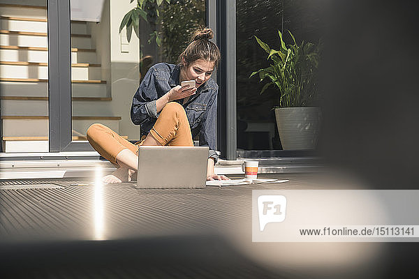 Junge Frau sitzt zu Hause auf der Terrasse und benutzt Handy und Laptop