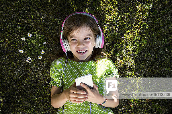Portrait of little girl lying on meadow listening music with headphones and smartphone