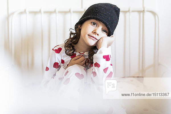 Porträt eines kleinen Mädchens  das mit Mütze und Pyjama im Bett sitzt