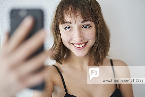 Porträt einer lächelnden jungen Frau  die sich zu Hause mit einem Smartphone selbstständig macht