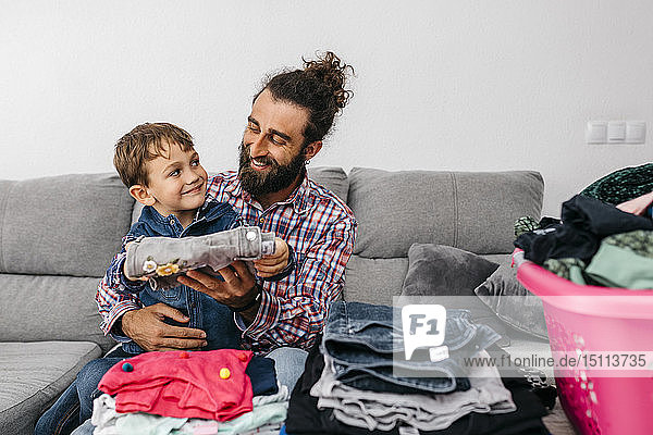 Porträt eines glücklichen Vaters und Sohnes  die zusammen auf der Couch sitzen und Wäsche zusammenlegen