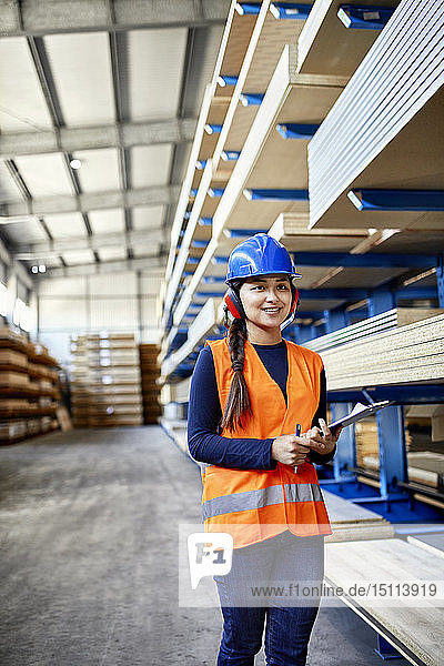 Lächelnde Arbeitnehmerin mit Ohrenschützern und Klemmbrett im Fabriklager