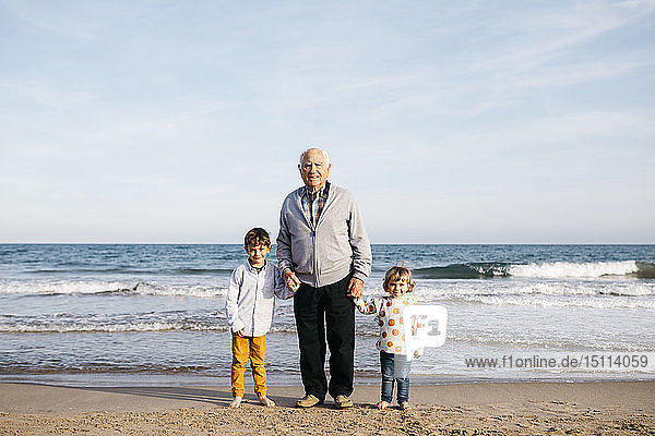 Porträt eines glücklichen Grossvaters  der mit seinen Enkelkindern Hand in Hand am Strand steht