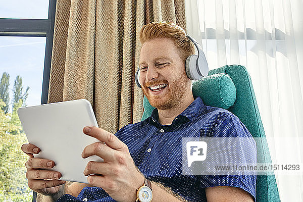 Junger Mann sitzt im Sessel  hört Musik und benutzt ein digitales Tablet