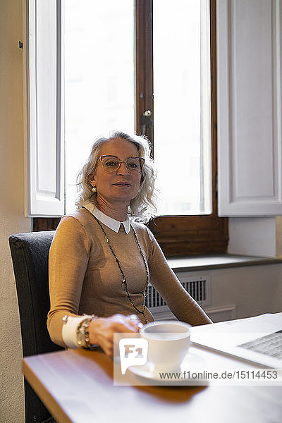 Porträt einer zufriedenen reifen Geschäftsfrau  die mit Laptop und einer Tasse Kaffee am Tisch sitzt