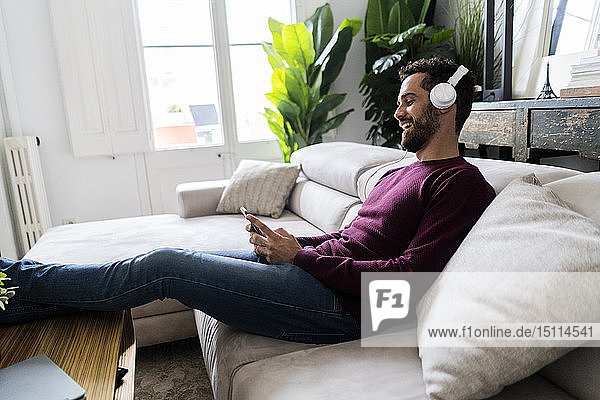 Lachender Mann auf Couch sitzend mit Handy und Kopfhörern