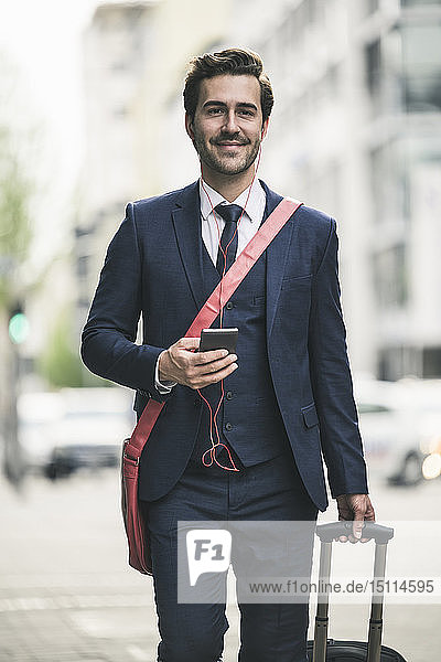 Confident businessman walking in the city with cell phone and suitcase