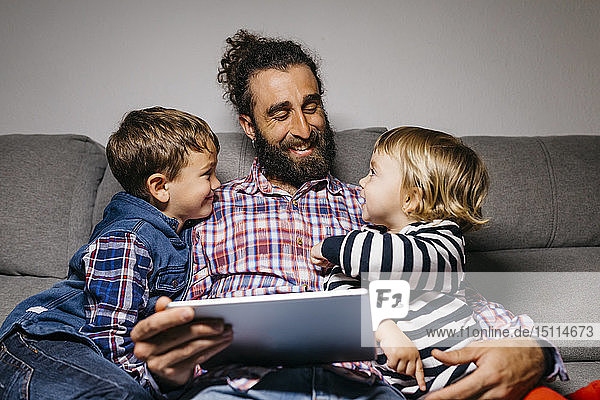 Glücklicher Vater sitzt mit seinen Kindern auf der Couch und benutzt digitales Tablet
