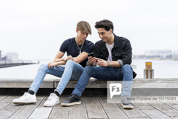 Denmark  Copenhagen  two young men sitting at the waterfront using cell phone