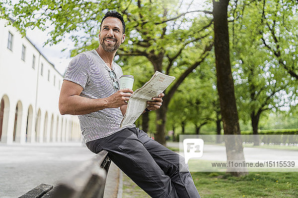 Lächelnder Mann mit Kaffee und Zeitung zum Mitnehmen auf der Parkbank