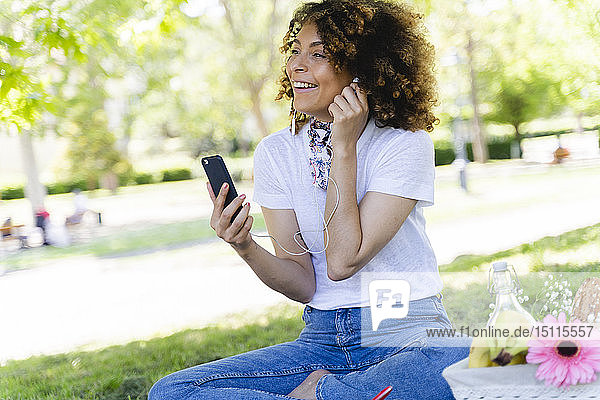 Glückliche Frau mit Handy und Kopfhörern beim Picknick im Park