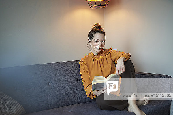 Porträt einer lächelnden Frau mit Buch  die zu Hause auf der Couch sitzt