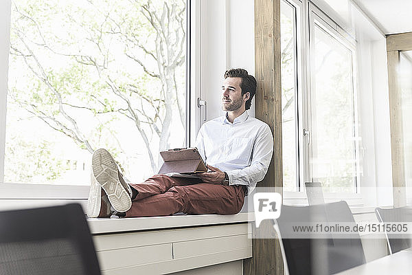 Junger Geschäftsmann sitzt auf der Fensterbank und benutzt ein digitales Tablett