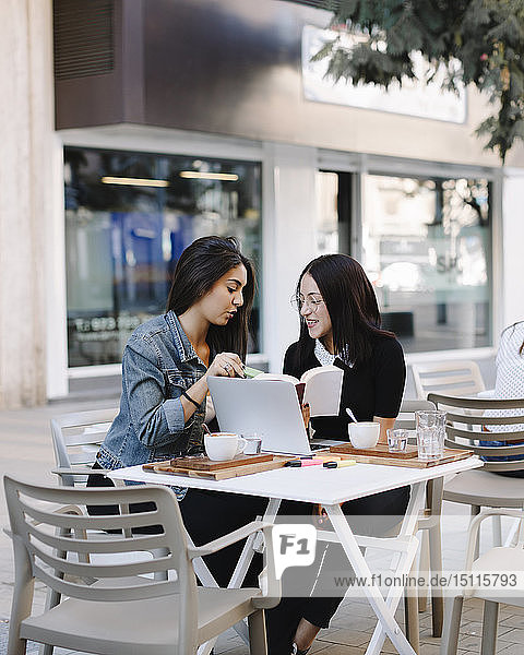 Two friends sitting together at a pavement cafe with book and laptop