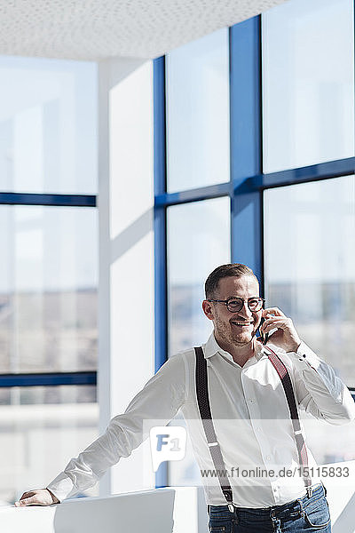 Glücklicher Geschäftsmann telefoniert am Fenster im modernen Büro mit seinem Handy