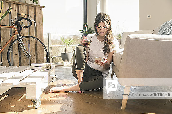 Young woman sitting on the floor at home with drink and cell phone
