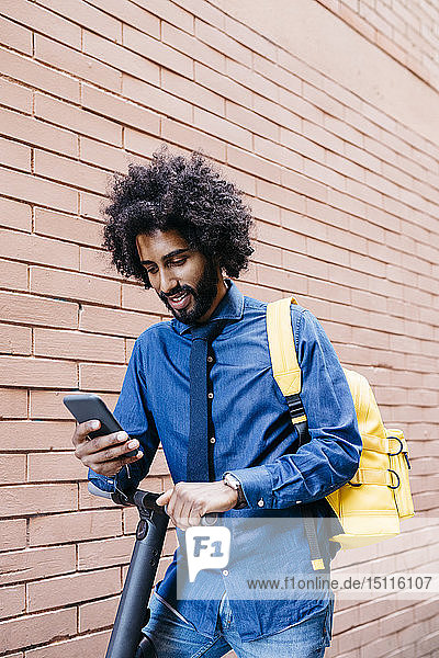 Porträt eines lächelnden jungen Mannes mit Rucksack und E-Scooter beim Blick auf das Handy vor der Ziegelmauer