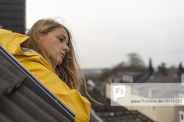 Nachdenkliche junge Frau im Regenmantel lehnt aus dem Dachfenster