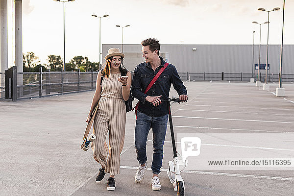 Happy young couple with cell phone  longboard and electric scooter on parking deck
