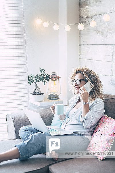 Frau auf Couch zu Hause mit Kaffeetasse  Laptop und Mobiltelefon