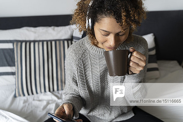 Woman sitting on bed  drinking coffee  listening music with headphones and smartphone