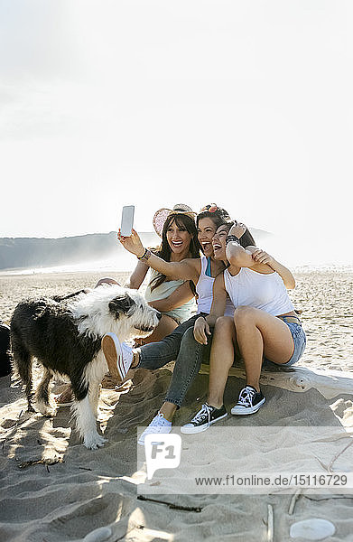 Glückliche Freundinnen mit Hund  die am Strand ein Selfie machen