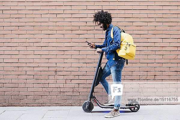 Junger Mann mit Rucksack und E-Scooter steht vor einer Ziegelmauer und schaut auf sein Handy
