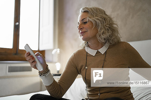 Zufriedene reife Geschäftsfrau sitzt am Bett und benutzt ein Mobiltelefon