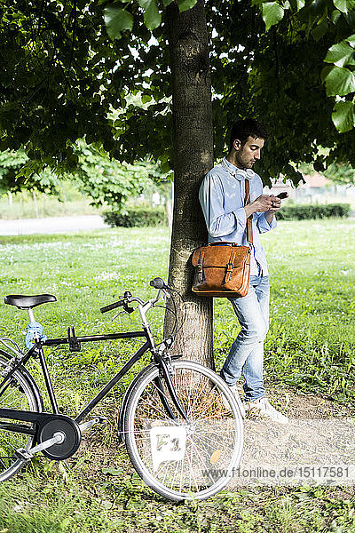 Young man with bicycle using smartphone  headphones around neck  standing under tree