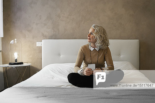 Reife Geschäftsfrau sitzt auf dem Bett mit einem Mobiltelefon  das aus dem Fenster schaut