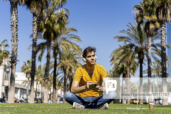 Spanien  Barcelona  Mann sitzt auf dem Rasen in der Stadt mit Kopfhörern und Notebook